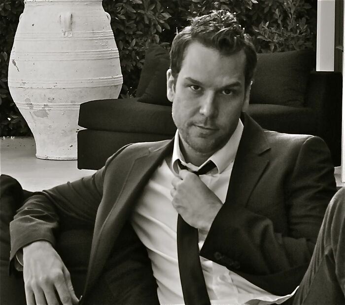 Dane Cook in a black suit and tie, sitting casually indoors with a confident expression and relaxed posture.