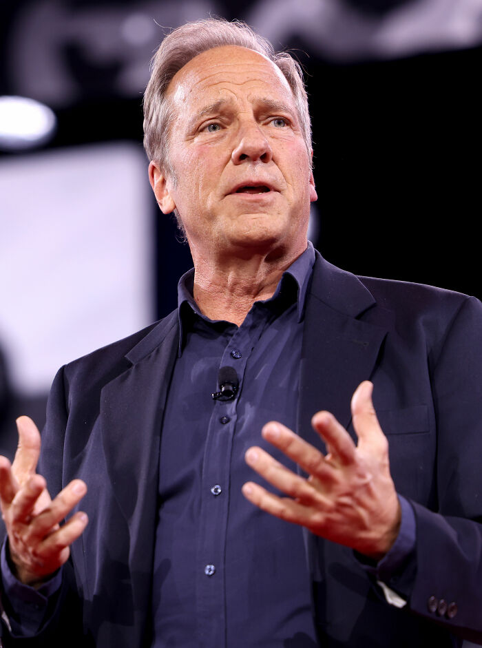 Mike Rowe speaking on stage in a dark blazer and shirt, highlighting key moments from his bio and career.