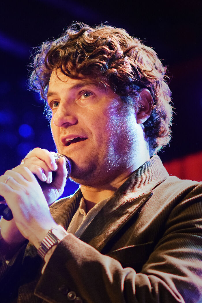 Adam Pally speaking into a microphone on stage, wearing a brown jacket under bright performance lighting.