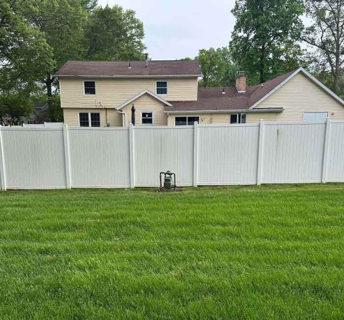 Suburban house behind a tall white fence with a well-maintained green lawn and trees in the background.