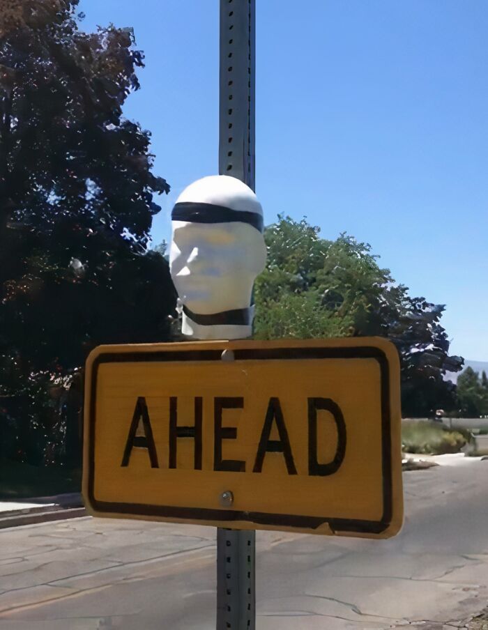 Mild vandalism on a street sign with a mannequin head wearing a black band covering eyes, placed above an ahead warning sign.