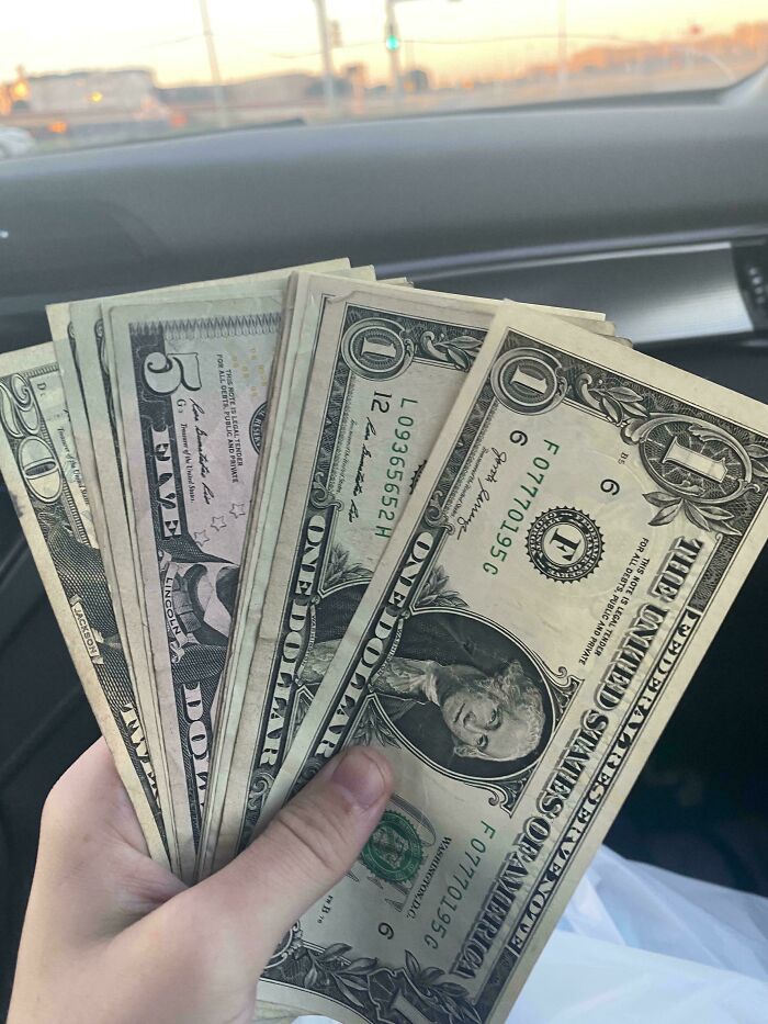 Hand holding various US dollar bills inside a car, a strange image that may linger in your brain longer than you want.