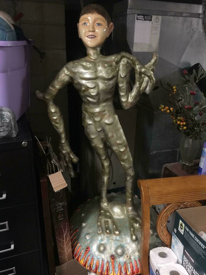 Weird and unexplainable sculpture of a humanoid figure with elongated limbs standing on a textured base indoors.