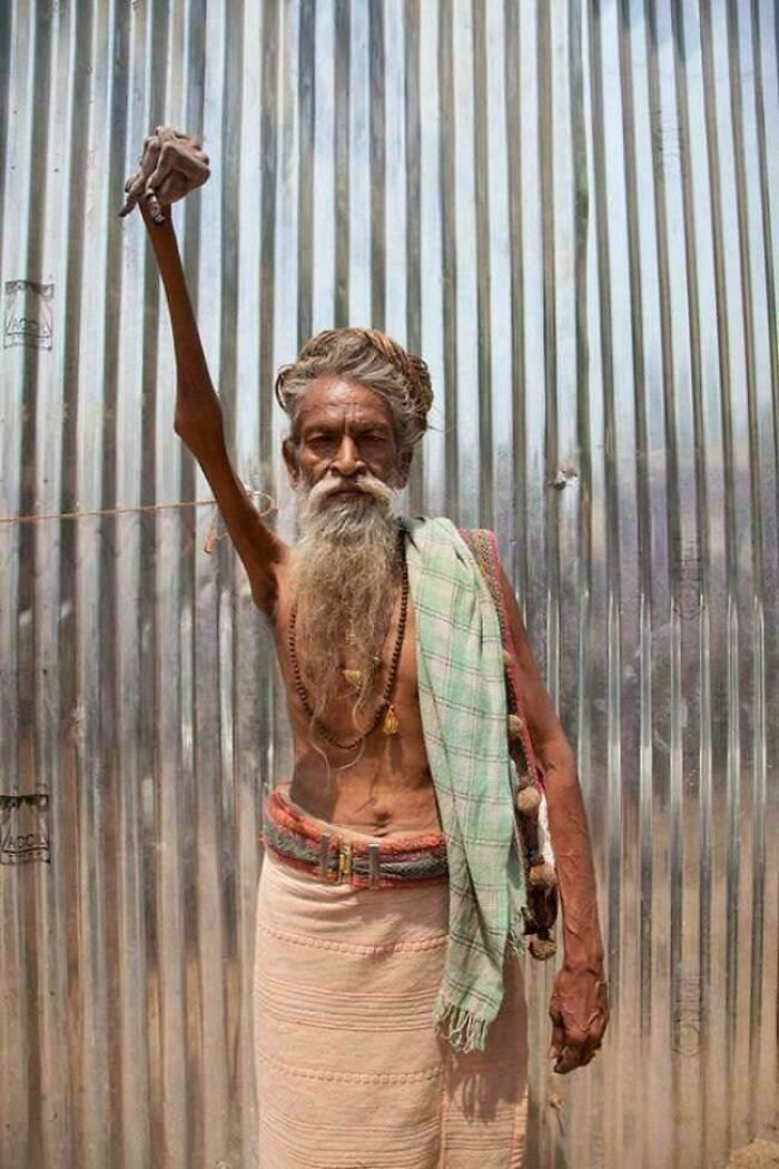 Elderly man with an exceptionally long and thin raised arm against a metal corrugated background, weird unexplainable image.