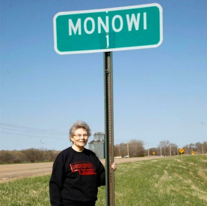 Elderly woman standing beside Monowi town sign, the only resident managing multiple roles in this strange earth location.