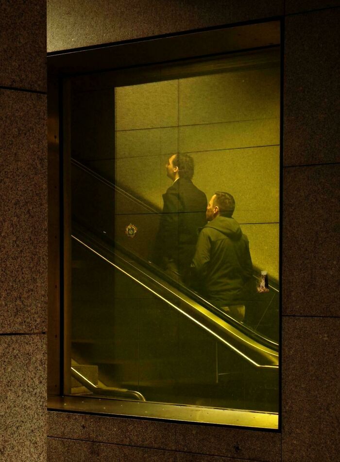 A reflection of men on an escalator in a yellow-lit window, showcasing an Accidental Renaissance look.