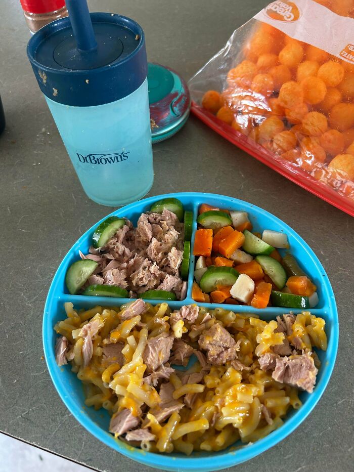 Blue divided plate with tuna, cucumbers, cheesy pasta, and mixed vegetables, next to a Dr. Brown's cup and a bag of cheese balls.