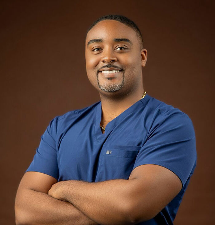 Plastic surgeon wearing blue scrubs smiling confidently with arms crossed against a brown background.