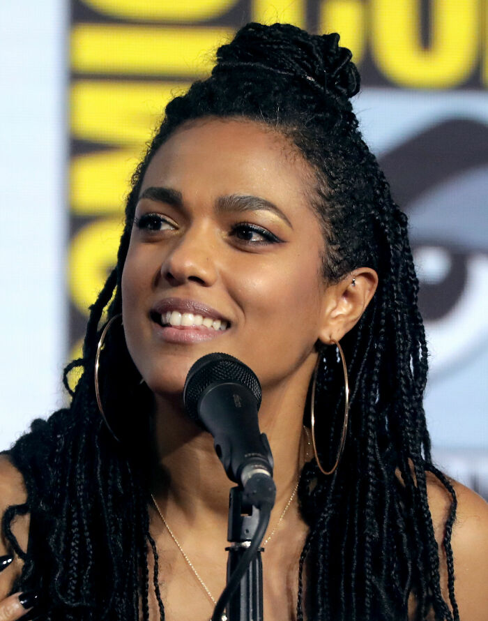 Freema Agyeman speaking at a panel event, wearing large hoop earrings and styled hair in a braided updo.