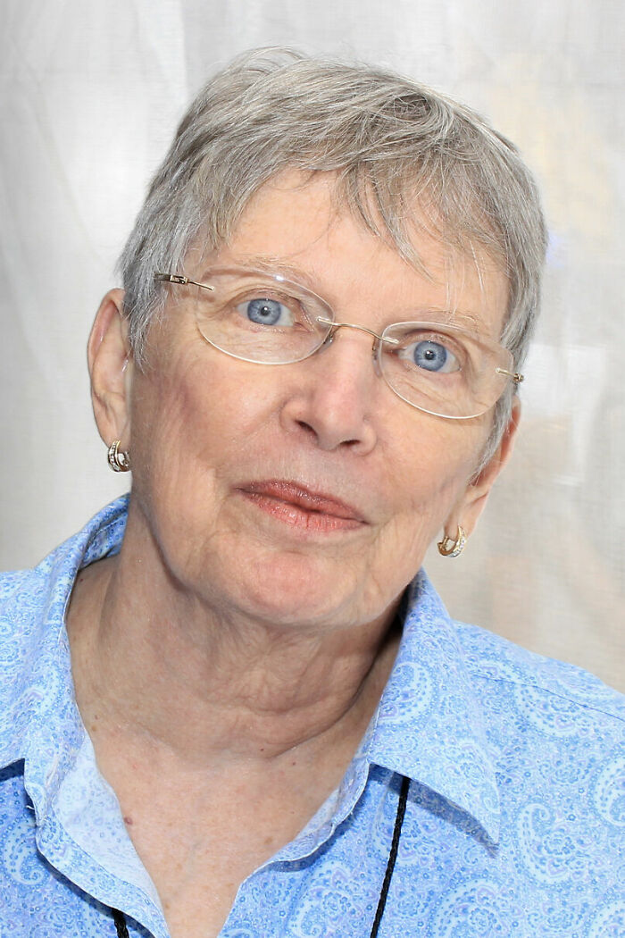 Lois Lowry with short gray hair and glasses, wearing a blue patterned shirt in a close-up portrait.