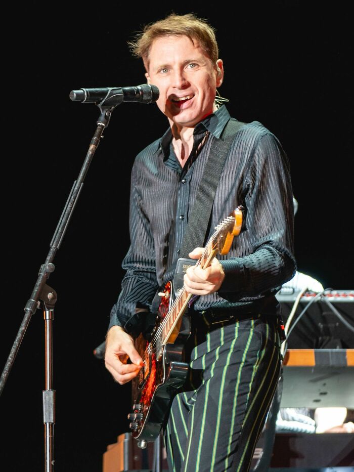 Alex Kapranos performing live on stage, playing guitar and singing into a microphone during a concert.