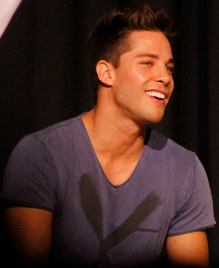 Dean Geyer smiling in a casual purple shirt with dark background, showcasing his bio and career highlights.