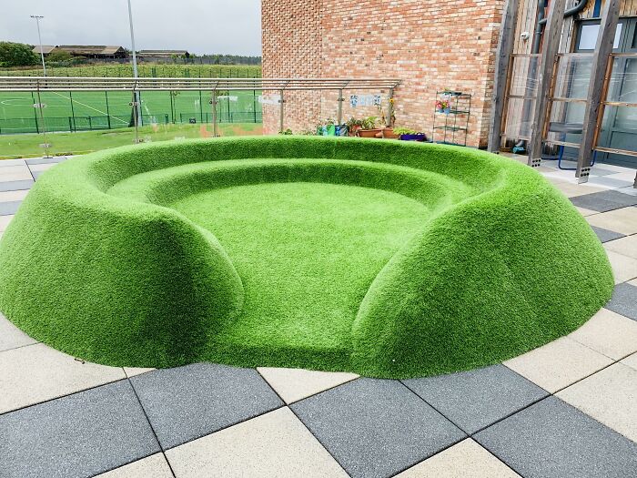Unedited photo of a green grass-like circular seating area outdoors on tiled ground near a brick building.