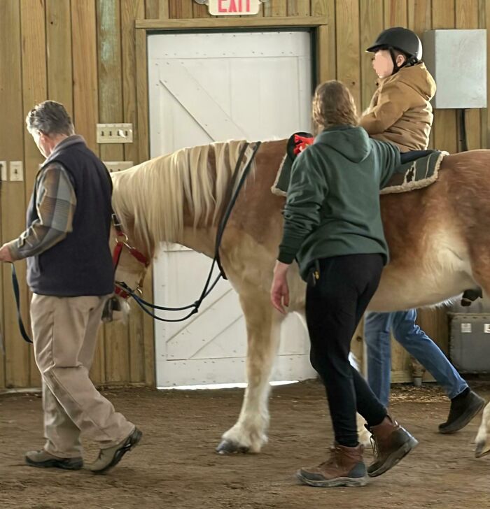 Child wearing helmet riding a horse indoors, guided by two adults in a setting that might mess with your already tired brain.
