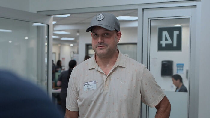 Man wearing a gray cap and visitor badge standing in a hospital hallway from The Pitt Season 2 guest star scene.