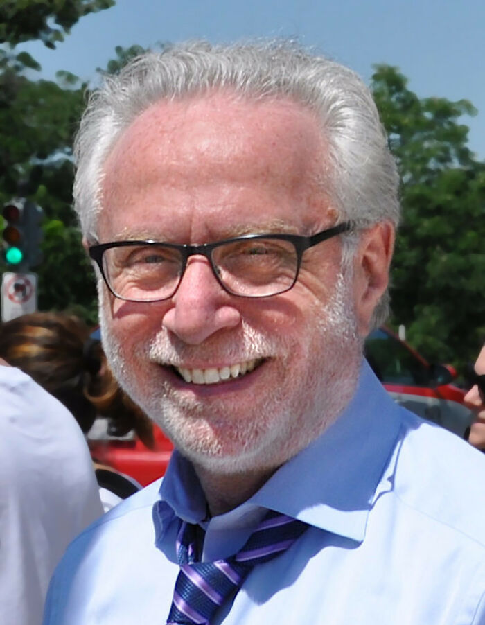 Wolf Blitzer smiling outdoors, wearing glasses, light blue shirt, and striped tie, with trees in the background.