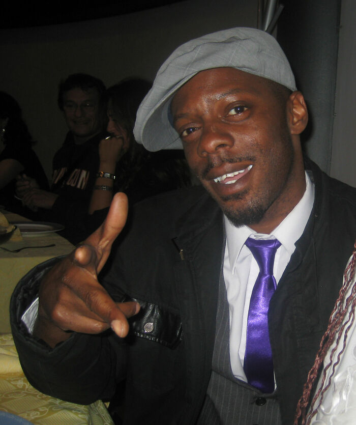 Ice MC wearing a gray cap and purple tie, smiling and gesturing with his hand in a casual indoor setting.
