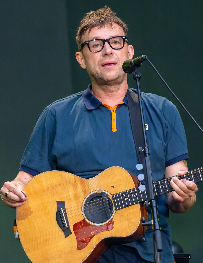 Damon Albarn performing live on stage, wearing glasses and playing acoustic guitar with microphone in front.