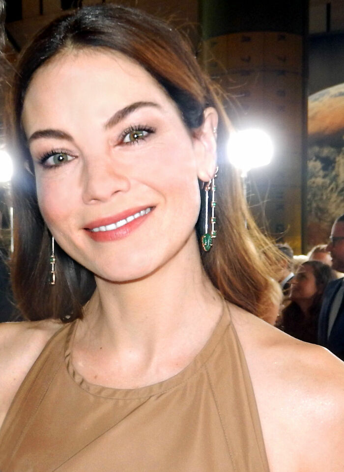 Michelle Monaghan smiling at an event, wearing dangling earrings and a brown halter top in a well-lit setting.