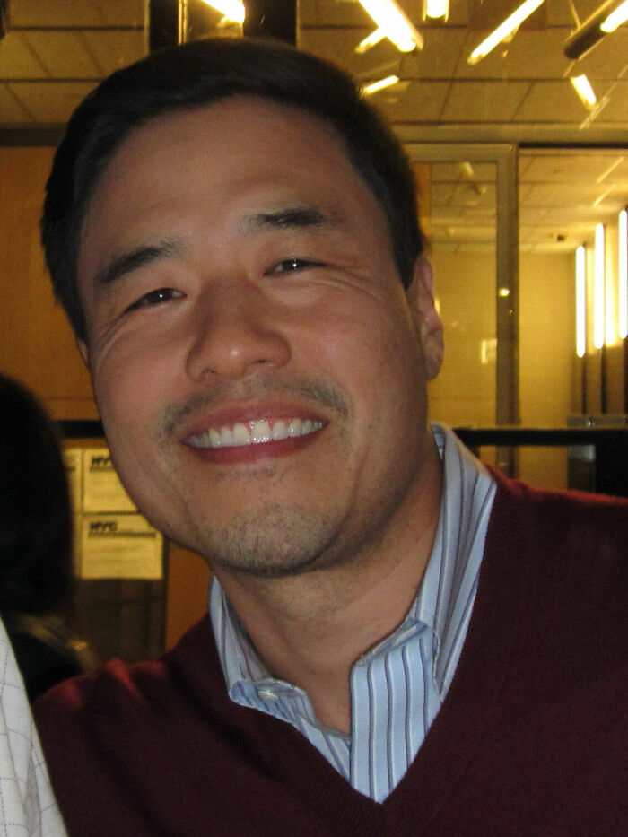 Randall Park smiling in a casual setting wearing a maroon sweater and striped shirt, highlighting his bio and career.