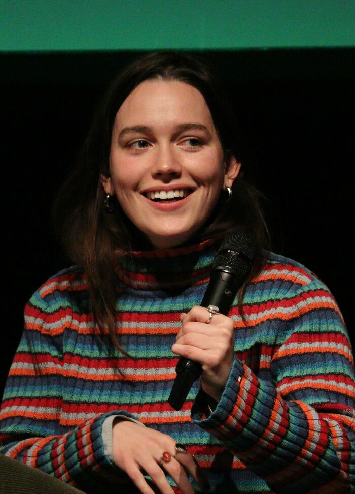 Victoria Pedretti smiling and speaking into a microphone, wearing a colorful striped sweater in a casual setting.