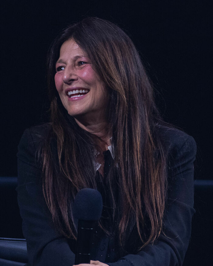 Catherine Keener smiling and holding a microphone during a speaking event on stage with a dark background.