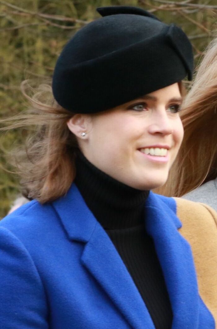 Princess Eugenie wearing a black beret and blue coat, smiling outdoors with a background of trees and natural light.