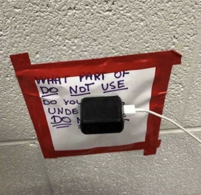 Charger plugged through a taped paper sign on wall showing first-world anarchist ignoring rules boldly.