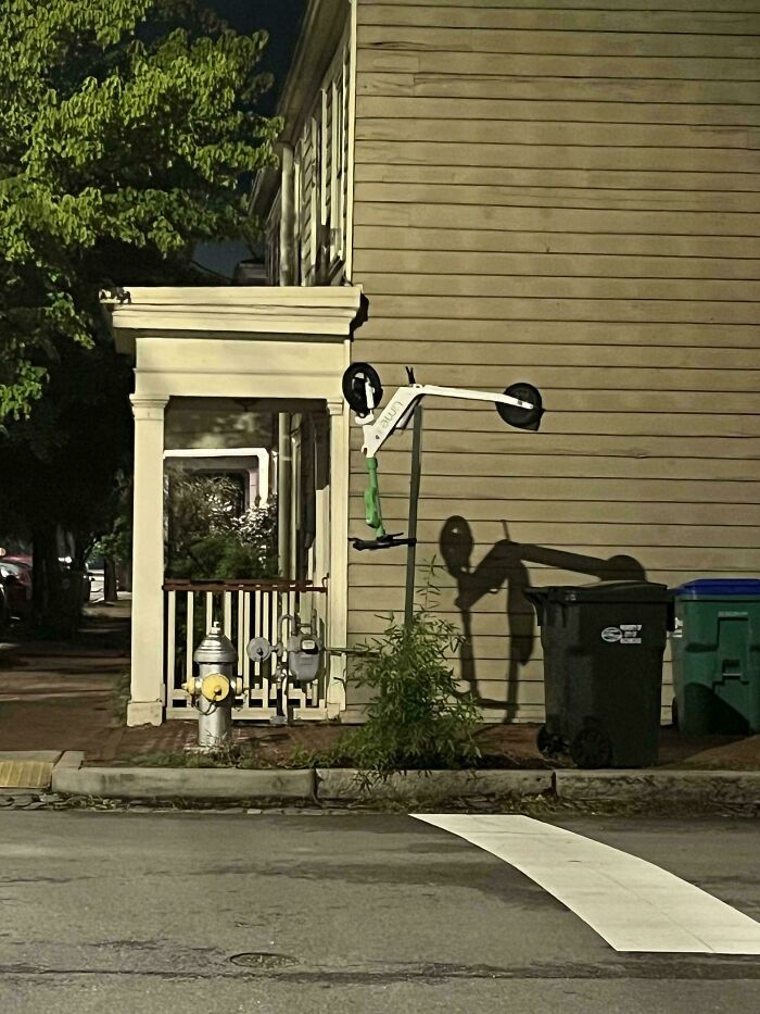 Electric scooter hanging upside down on pole casting a shadow near trash bins and a fire hydrant at night, ungovernable anarchist style.