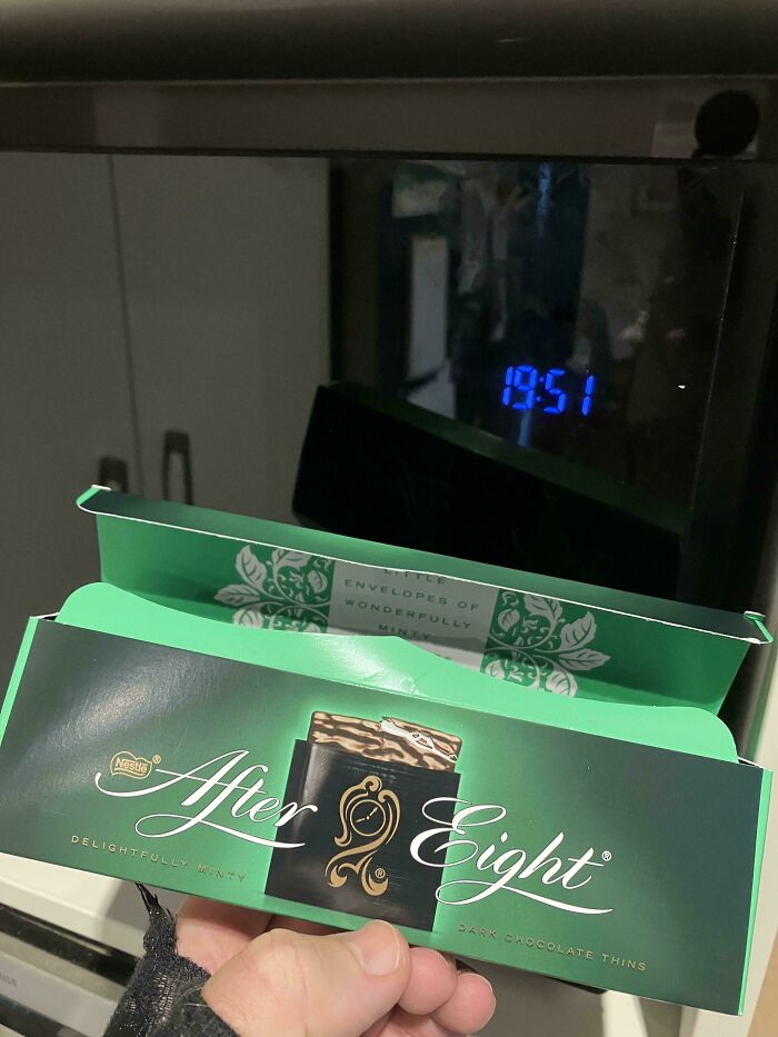 Box of After Eight dark chocolate thins being held open against a dark reflective surface showing a digital clock at 19:51.