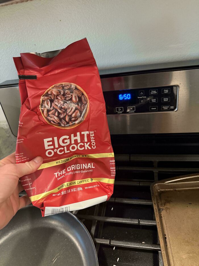 Hand holding a red bag of Eight O’Clock coffee whole bean medium roast on a kitchen stove background.
