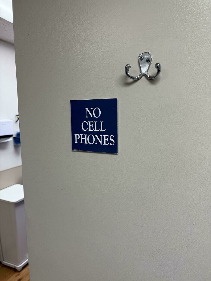 No cell phones sign posted on a plain wall near a silver dual hook in an indoor setting showing ungovernable rules.