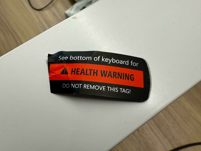 Health warning label on keyboard with bold text warning not to remove tag on a white surface and wooden floor background.