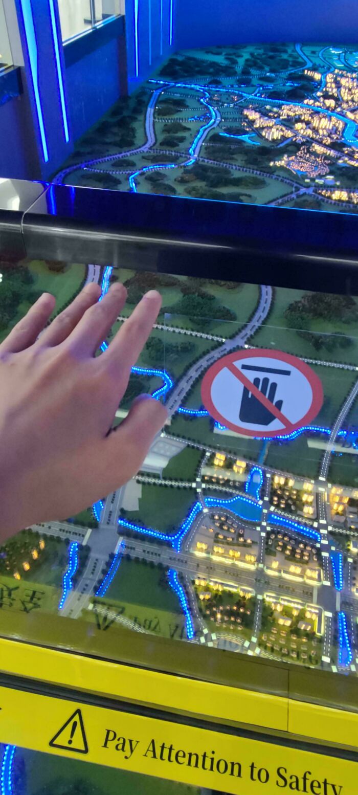 Hand reaching over a model city display with illuminated roads and a no-touch sign, reflecting first-world anarchist defiance.