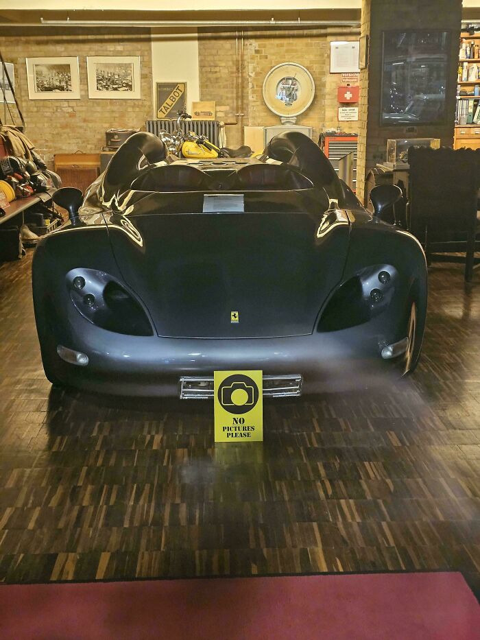 Black sports car displayed indoors with a no pictures sign, symbolizing first-world anarchists who challenge norms.