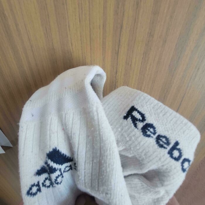 Two white socks with Adidas and Reebok logos displayed against a wooden background featuring first-world anarchists style.