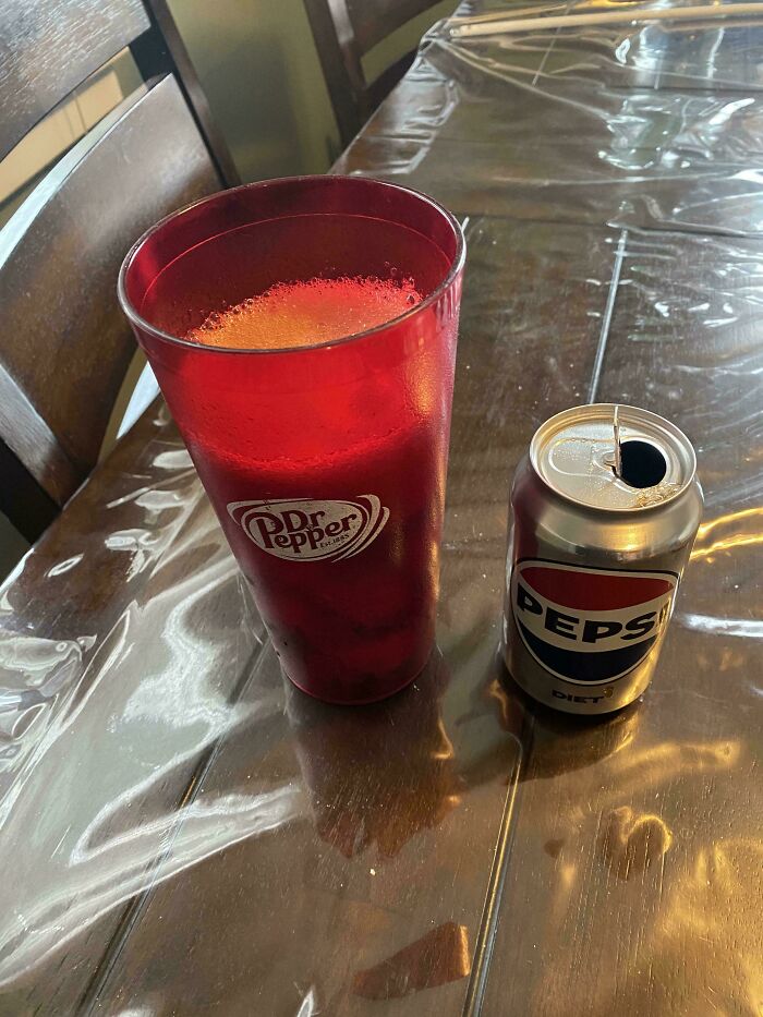 Red Dr Pepper cup filled with soda beside a small opened Pepsi can on a wooden table reflecting light.
