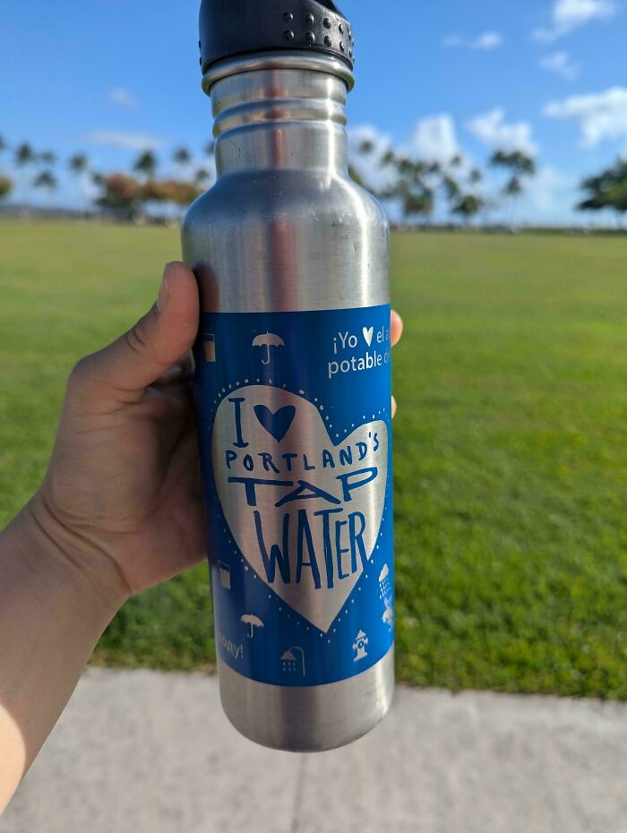 Hand holding a stainless steel water bottle with a Portland tap water sticker in a park under blue sky.