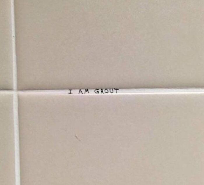 Small text graffiti on bathroom tile grout humorously stating I am grout, a mild vandalism example.