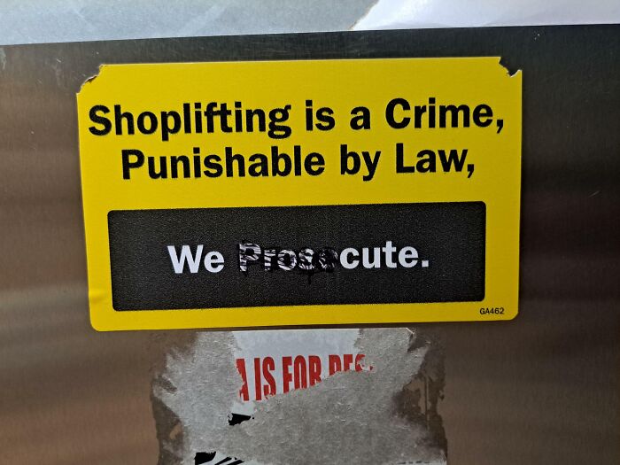 Yellow sign warning that shoplifting is a crime with mild vandalism partially obscuring the word prosecute.