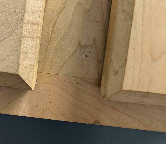 Tiny mild vandalism drawing of a cat face on wooden surface underneath stacked blocks, showing subtle creative mischief.