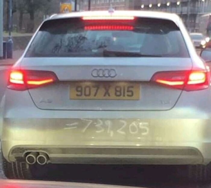Silver Audi car with mild vandalism showing a dirty rear bumper written with numbers on a city street.