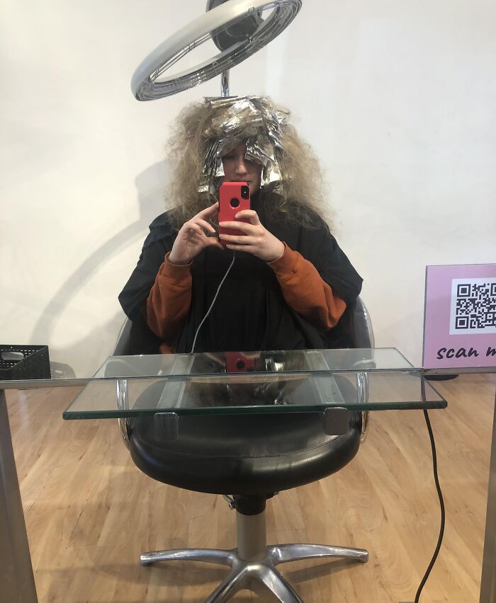 Person with foil in hair at salon, taking a selfie with a red phone in front of a mirror, unusual unedited photo.