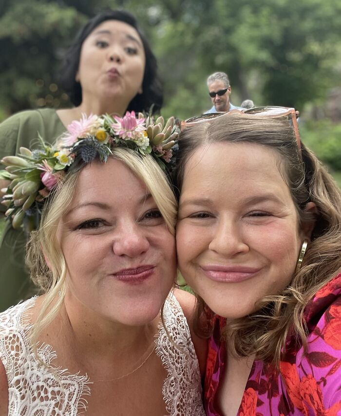 Two women smiling closely with flower crown and sunglasses, a photobombing woman making a funny face behind in unedited photo.