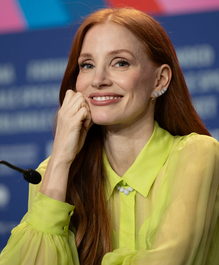 Jessica Chastain smiling at a press event, wearing a lime green blouse and delicate floral earrings.