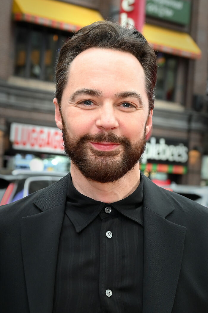 Jim Parsons with a beard wearing a black shirt and jacket, smiling outdoors in an urban setting for bio and career highlights.