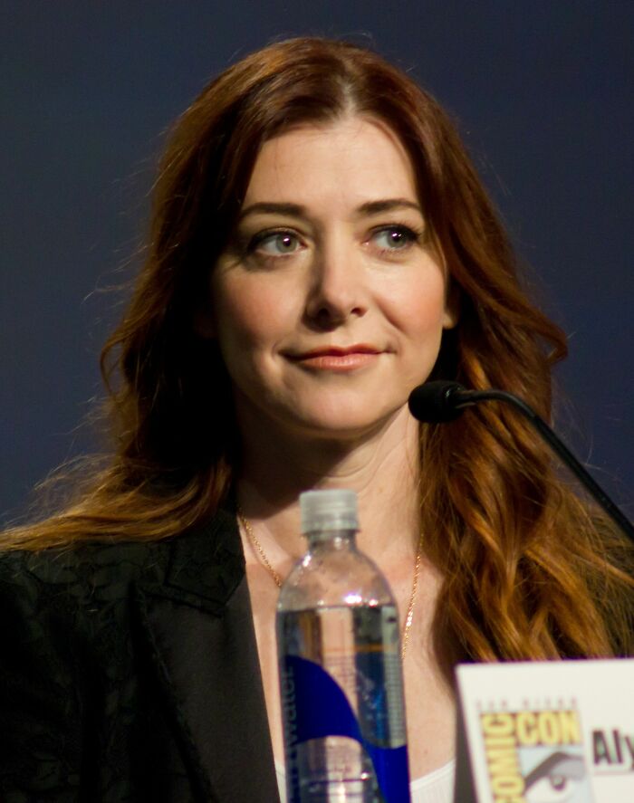 Alyson Hannigan speaking at a panel event, with long red hair and a microphone in front of her.