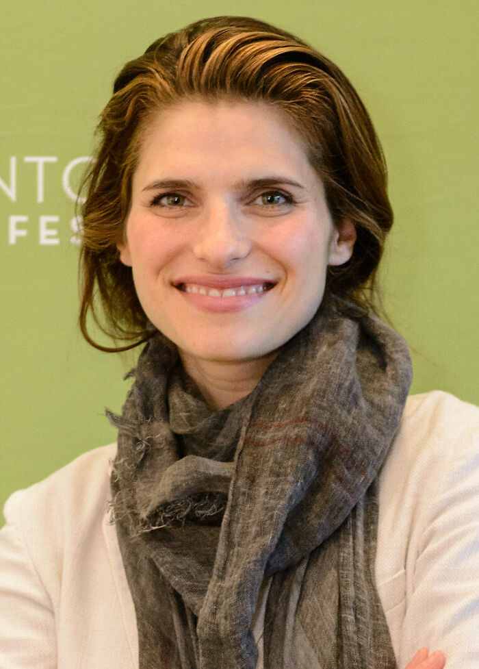 Lake Bell smiling and wearing a gray scarf and white top against a green background at a public event.