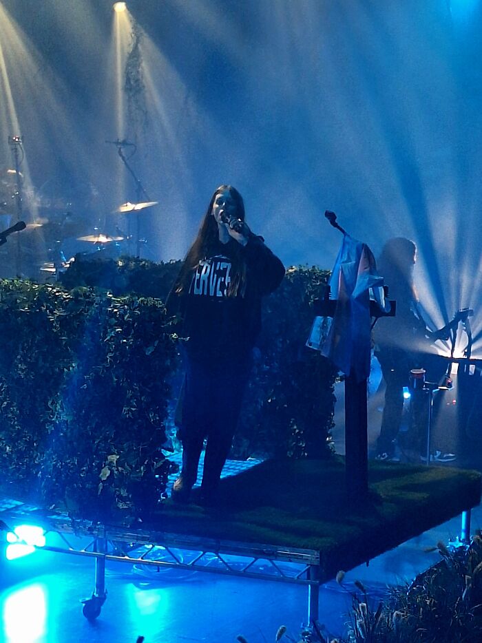 Ethel Cain performing on stage with moody blue lighting and surrounded by foliage during a live concert.
