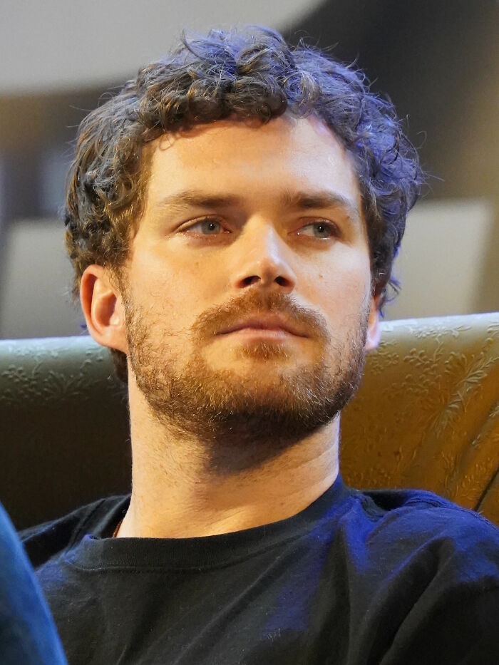 Finn Jones with curly hair and beard, wearing a dark shirt, sitting thoughtfully in a well-lit indoor setting.